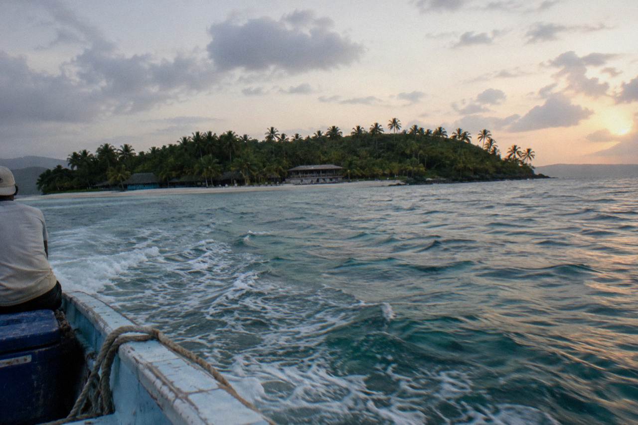 A small palm covered island with white sand and turquoise water seen across a calm bay