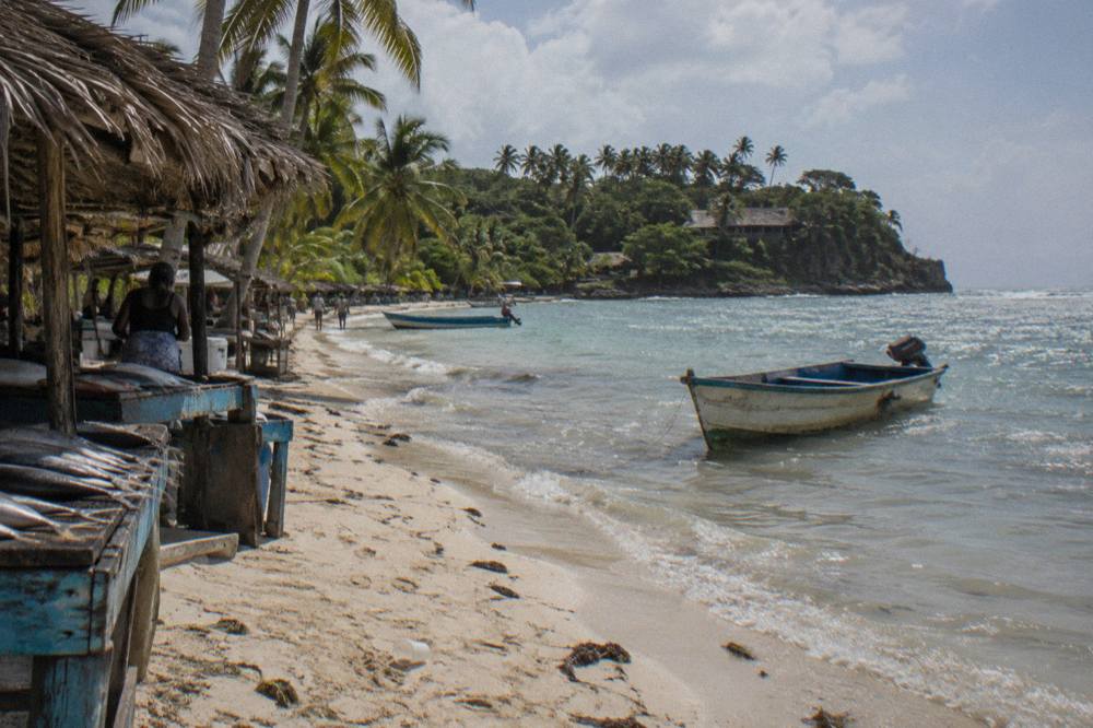 A white sand public beach with palm trees and calm shallow water, small thatched food stalls along the treeline