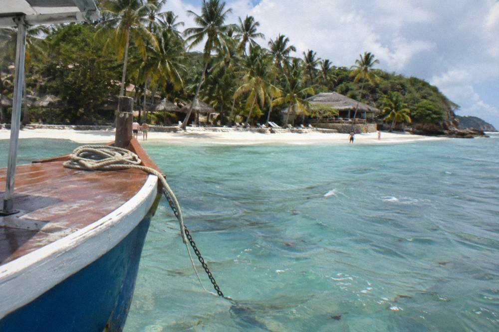 A boat anchored in clear shallow water beside a palm lined island beach in a wide blue bay