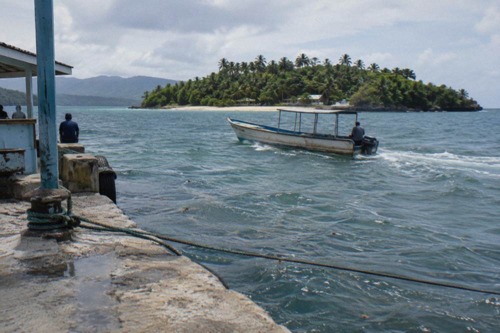 A wooden boat crossing calm turquoise bay water toward a green island with a white beach