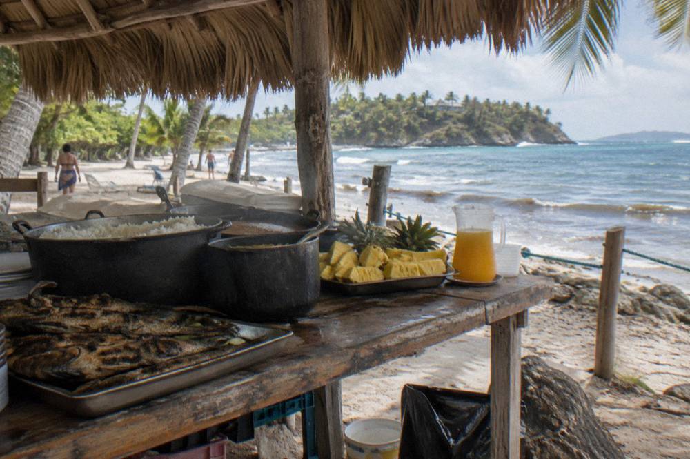 Beach buffet table with grilled fish, rice and tropical fruit under a thatched palm roof