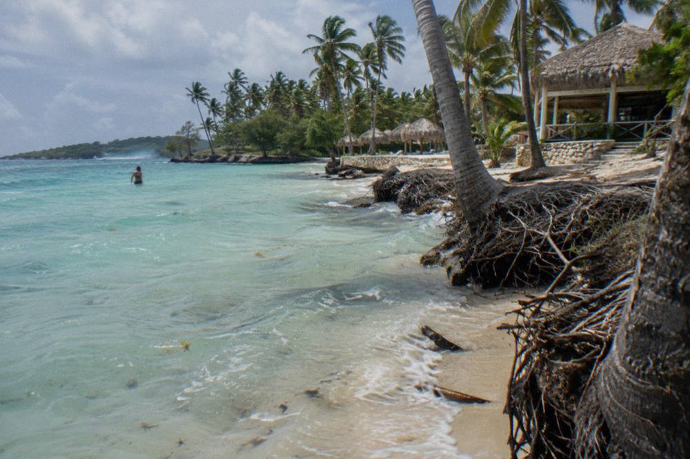 A white sand beach with leaning palm trees and shallow turquoise water along a small island shoreline