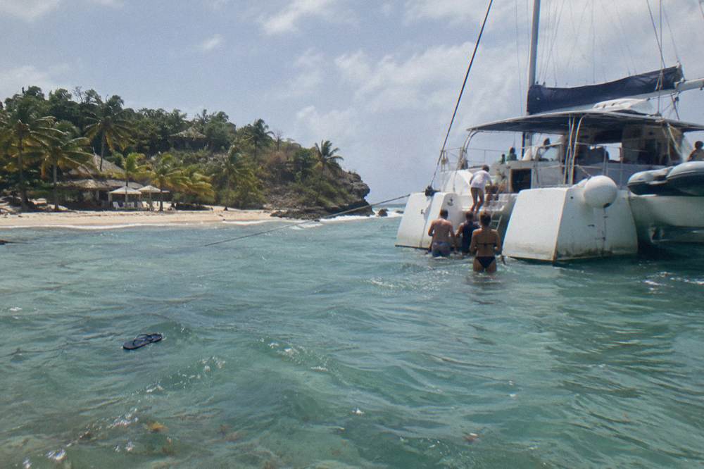 A catamaran anchored near an island beach with passengers wading in clear shallow water