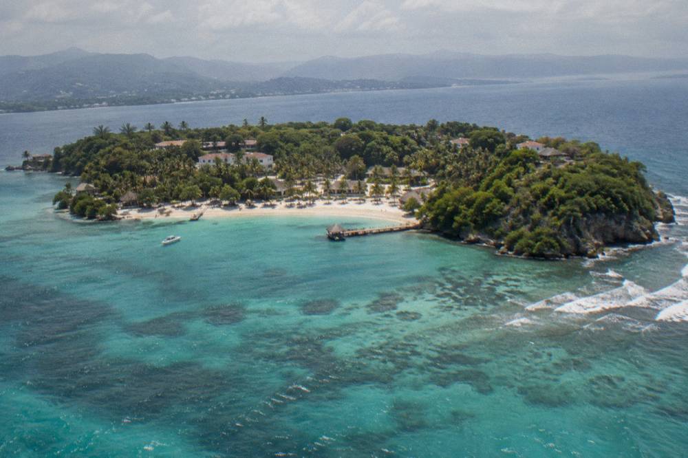 An aerial view of a small green island surrounded by turquoise bay water near a distant coastline
