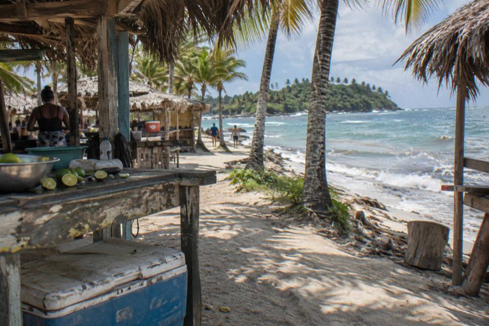 Simple beach food stalls and thatched shade structures behind a narrow sand path