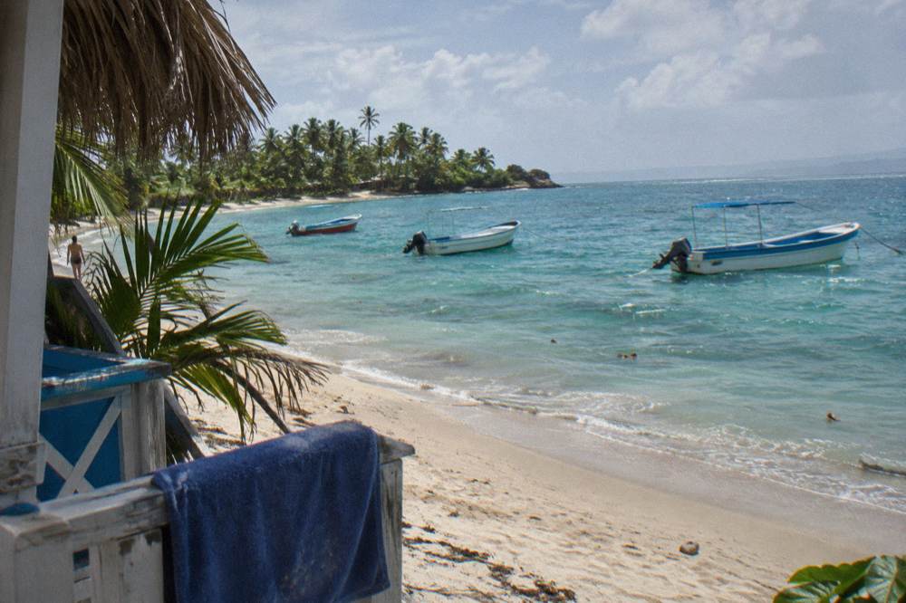 Small boats anchored offshore from a narrow palm-lined beach in late morning light