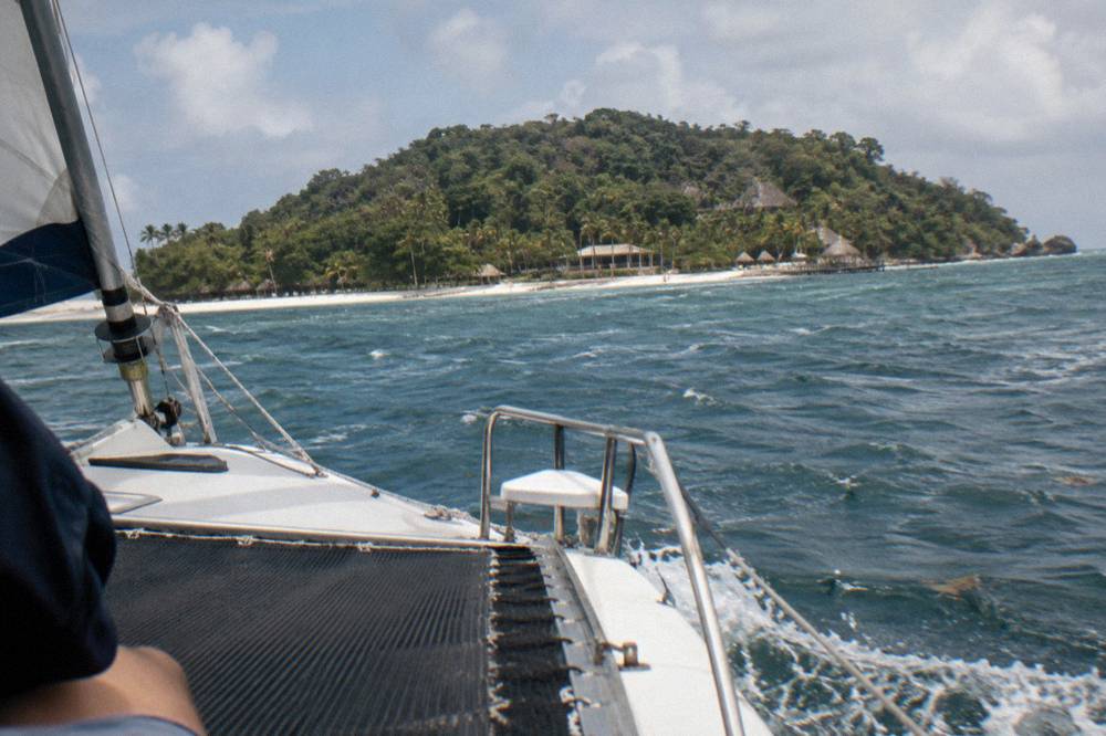 A catamaran cutting through light chop on blue bay water with hills in the background