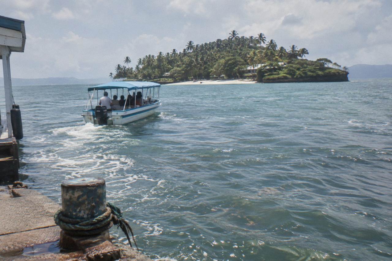 A small passenger boat crossing calm turquoise water toward a palm covered island in Samana Bay