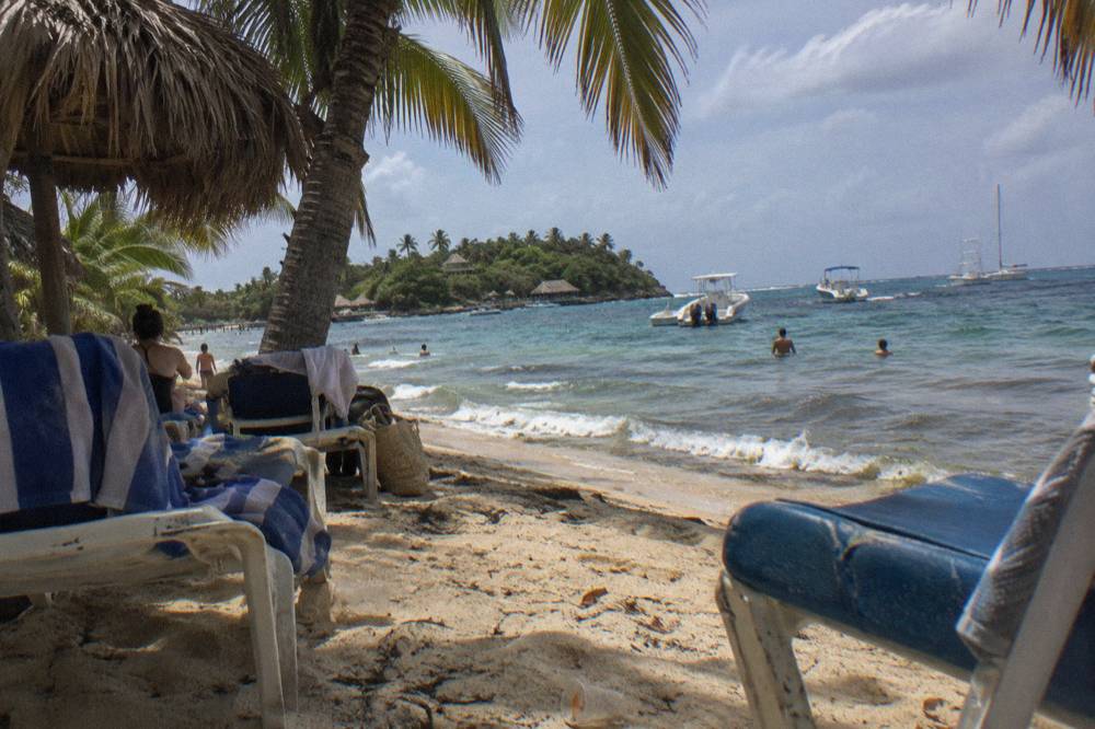 Shaded thatched umbrellas and loungers on a palm-lined beach with a few boats moored offshore