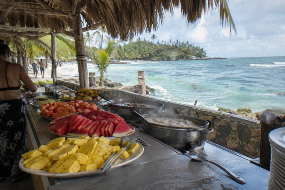 Beach buffet serving line with tropical fruit platters under a thatched roof