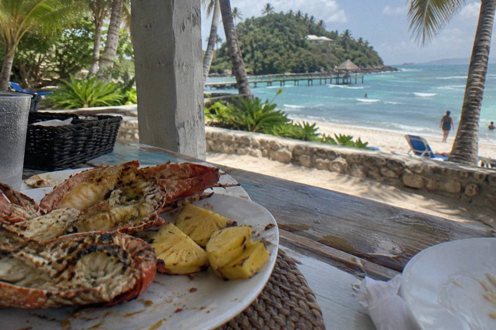A shaded beach buffet table with grilled lobster and tropical fruit beside turquoise water