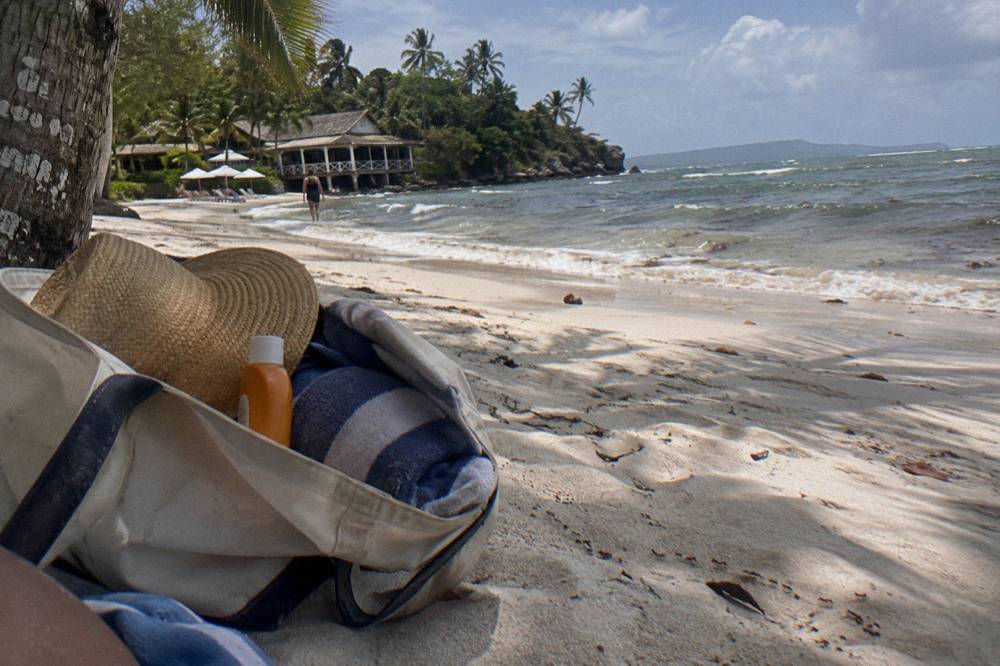 A beach bag with sunscreen, hat and towel on white sand under palm shade