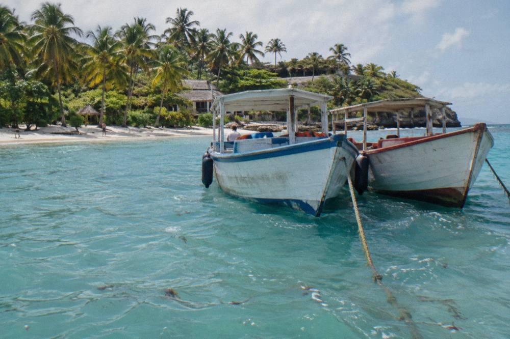 Wooden tour boats anchored in shallow turquoise water beside a palm-lined island beach