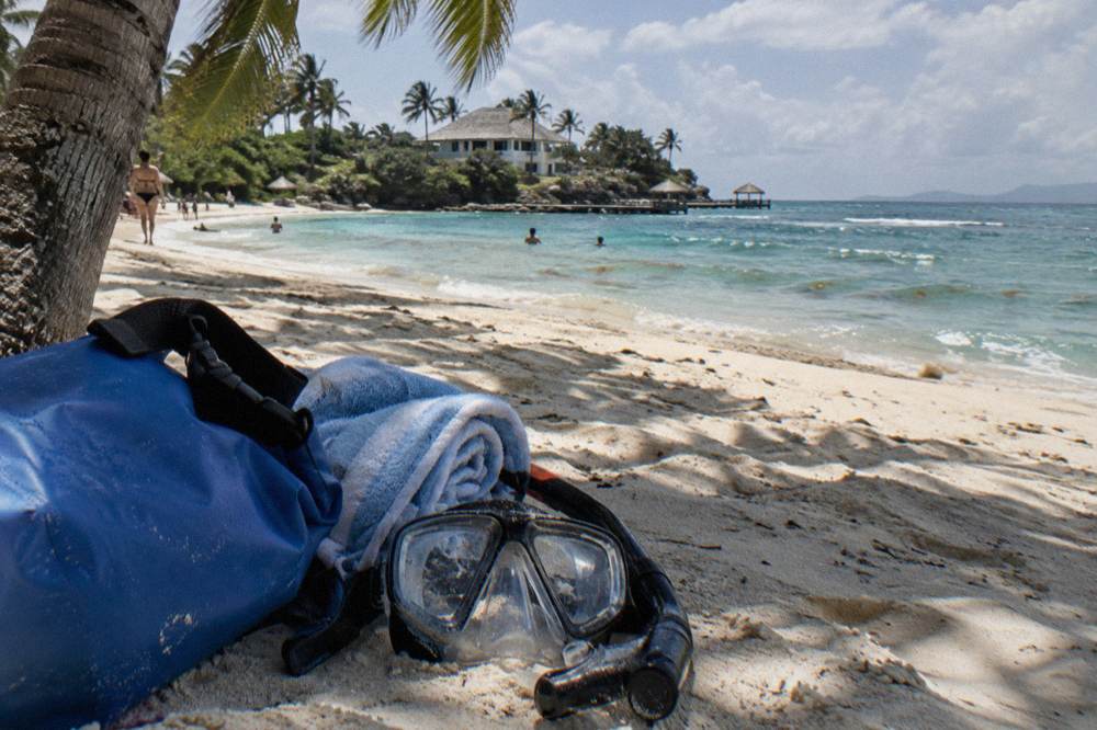 Snorkel mask and towel resting on sand under palm shade beside calm water