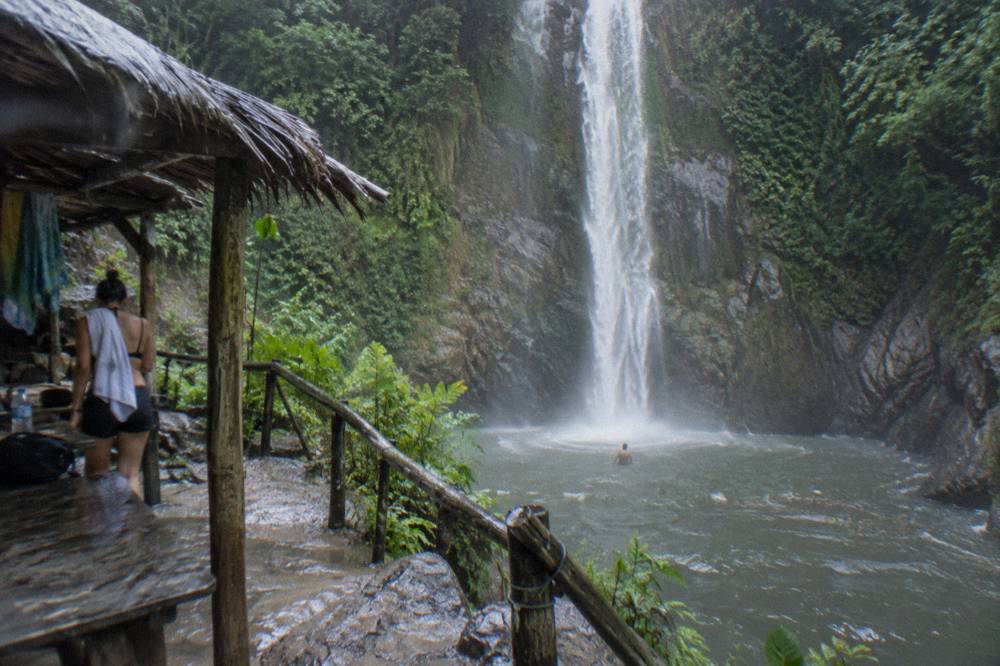 Tall waterfall framed by green cliffs with a swimmer standing in the mist below