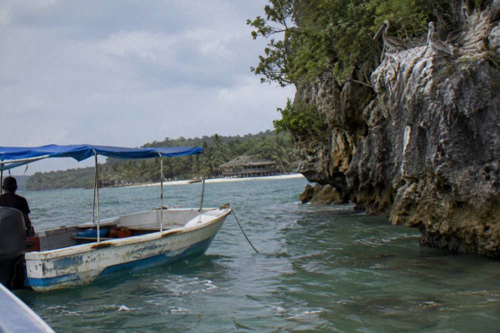Small boat anchored in shallow clear water beside a limestone cliff with pelicans perched above