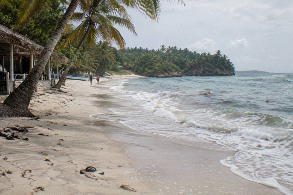 Long empty beach with white sand and palm trees curving toward green headlands