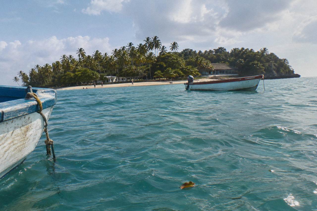 A calm turquoise bay with a small palm covered island and boats anchored offshore